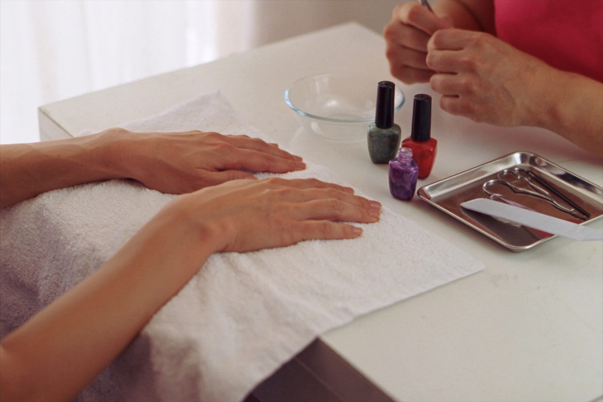 A client's hands resting on a folded white towel at a manicure table, with a small steel tray of tools, a glass bowl and three bottles of polish beside them, in warm daylight.